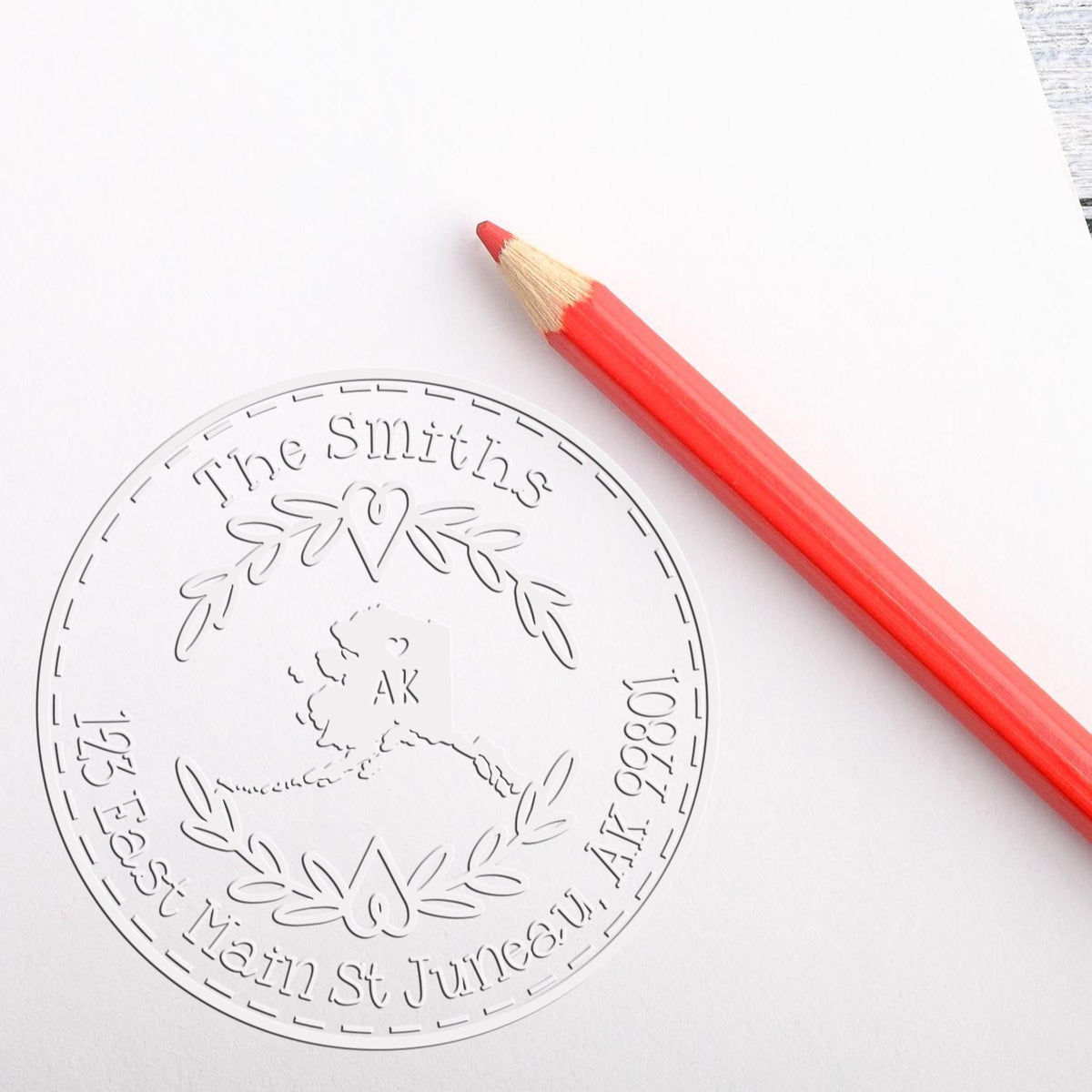 Clear round custom seal stamp with engraved wreath and AK logo beside a red pencil on white paper.