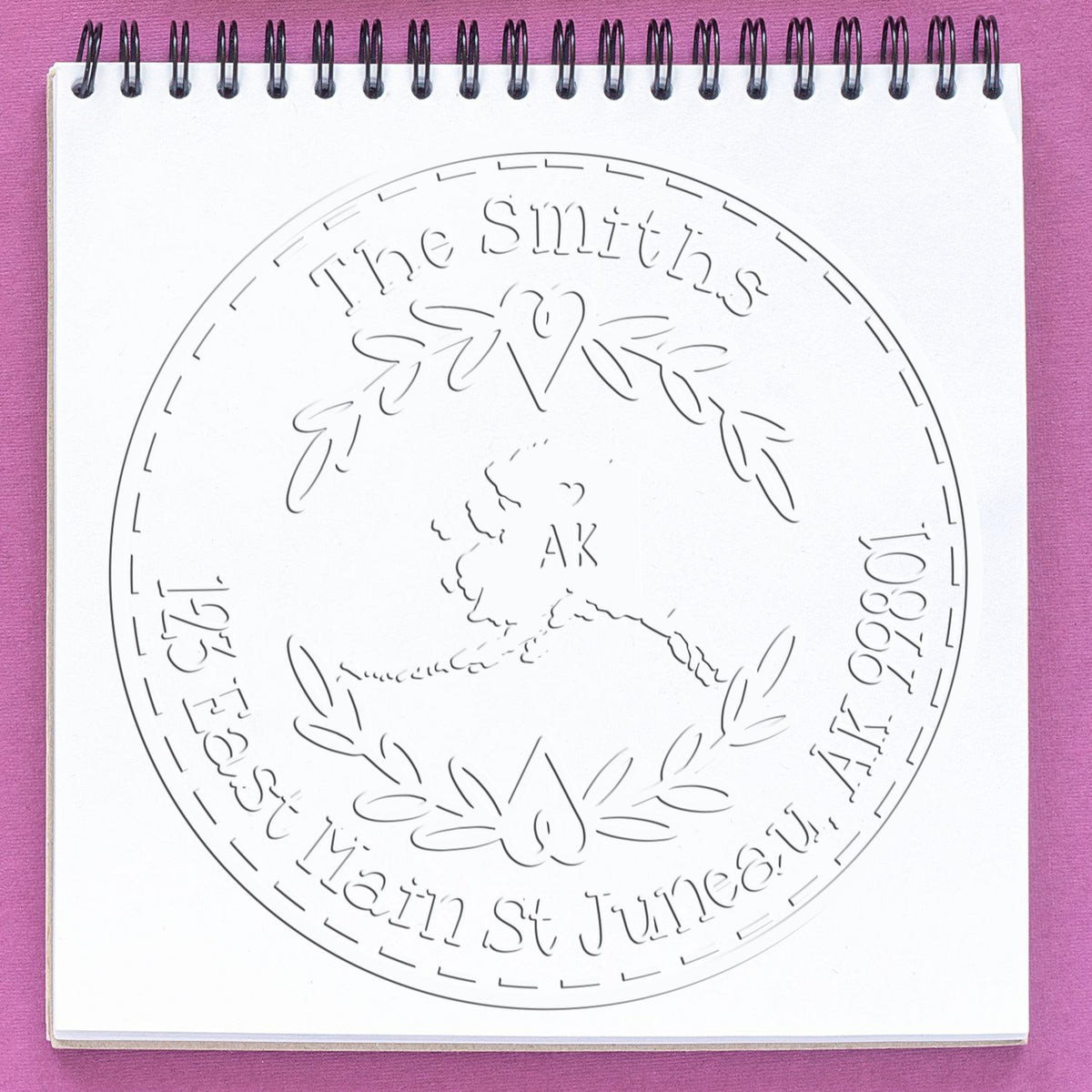 Embossed white circular stamp seal for The Smiths, with leaf wreath, heart and AK details on a spiral notebook, pink background.