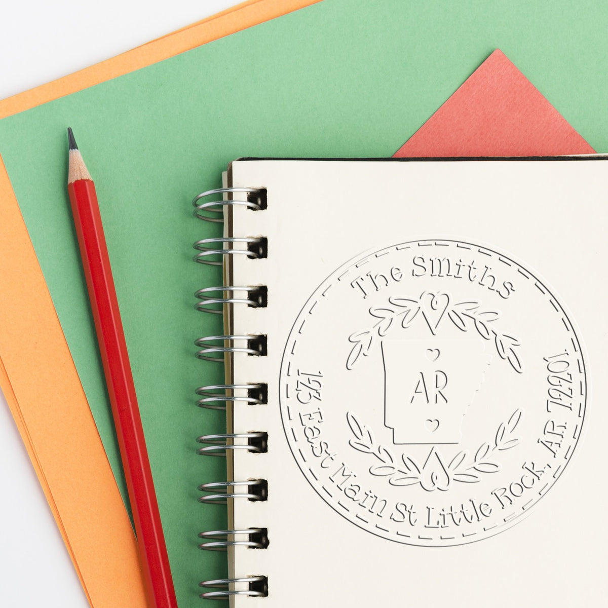 Custom stamp seal embossed on spiral notebook, with a red pencil and green, orange, and coral paper background.