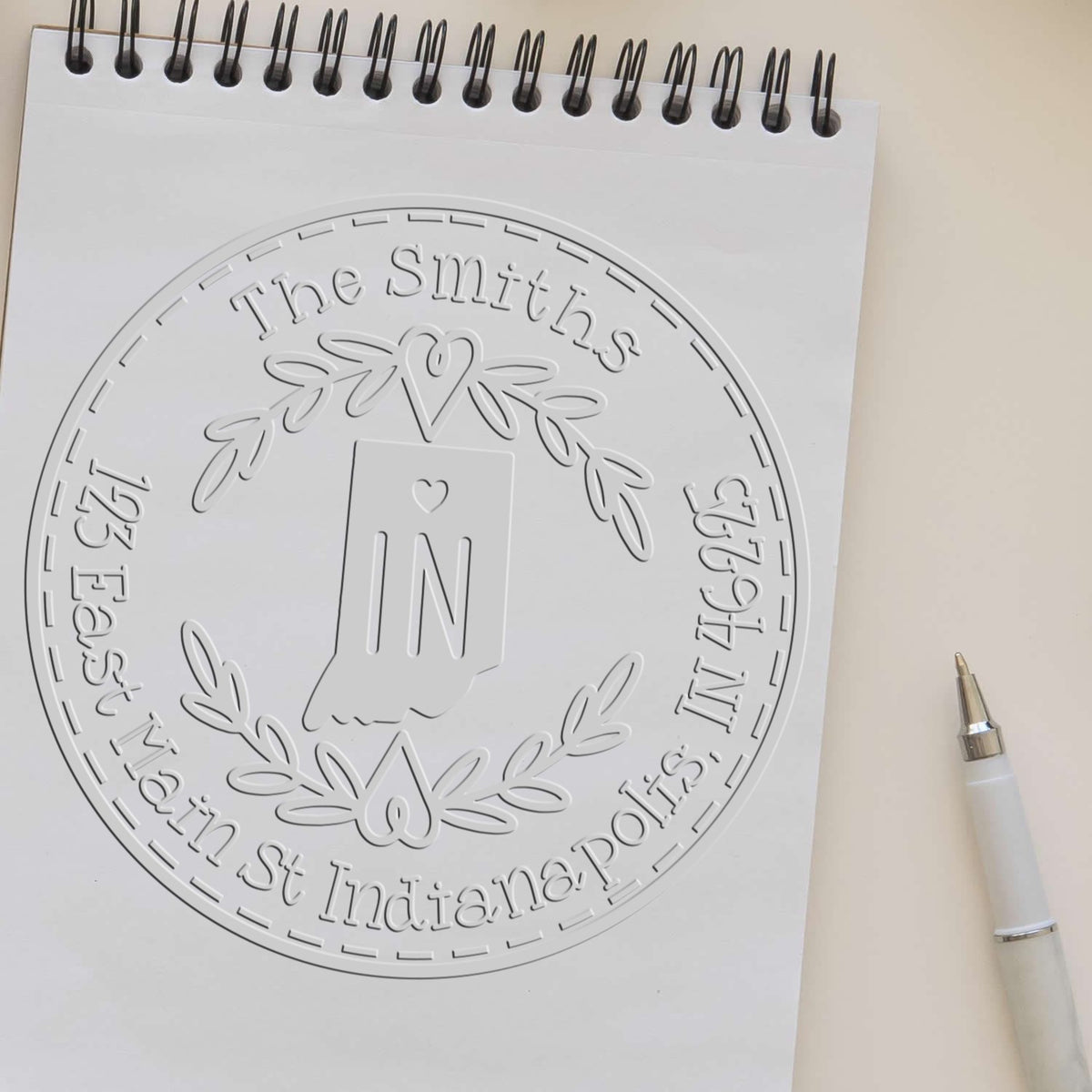 Embossed white round stamp seal on spiral notebook with The Smiths, Indiana, laurel wreath, and monogram N; silver pen beside it.
