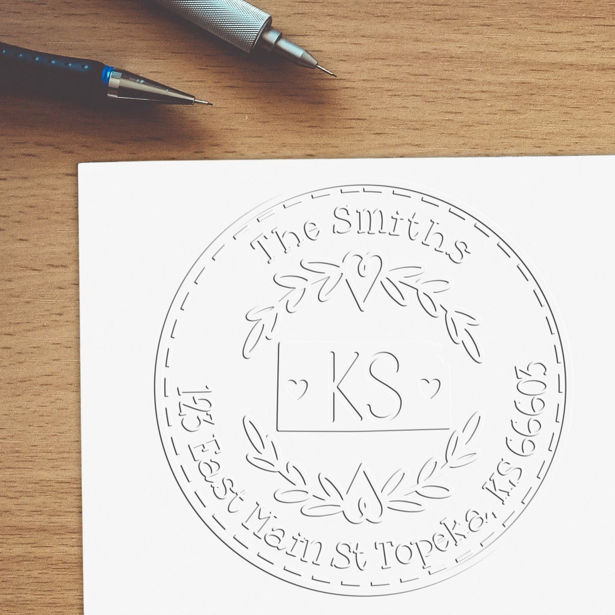 Custom white embossing stamp and seal design for The Smiths with KS monogram, heart and laurel wreath on wooden desk beside two pens.