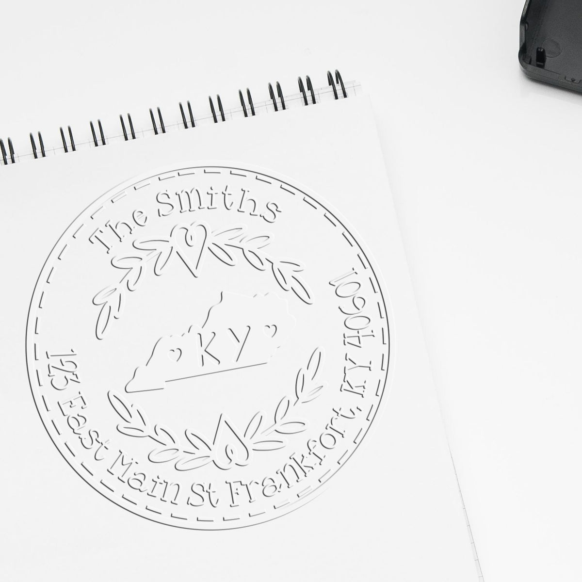 White spiral notebook with embossed circular custom stamp seal, floral wreath, and KY address detail.