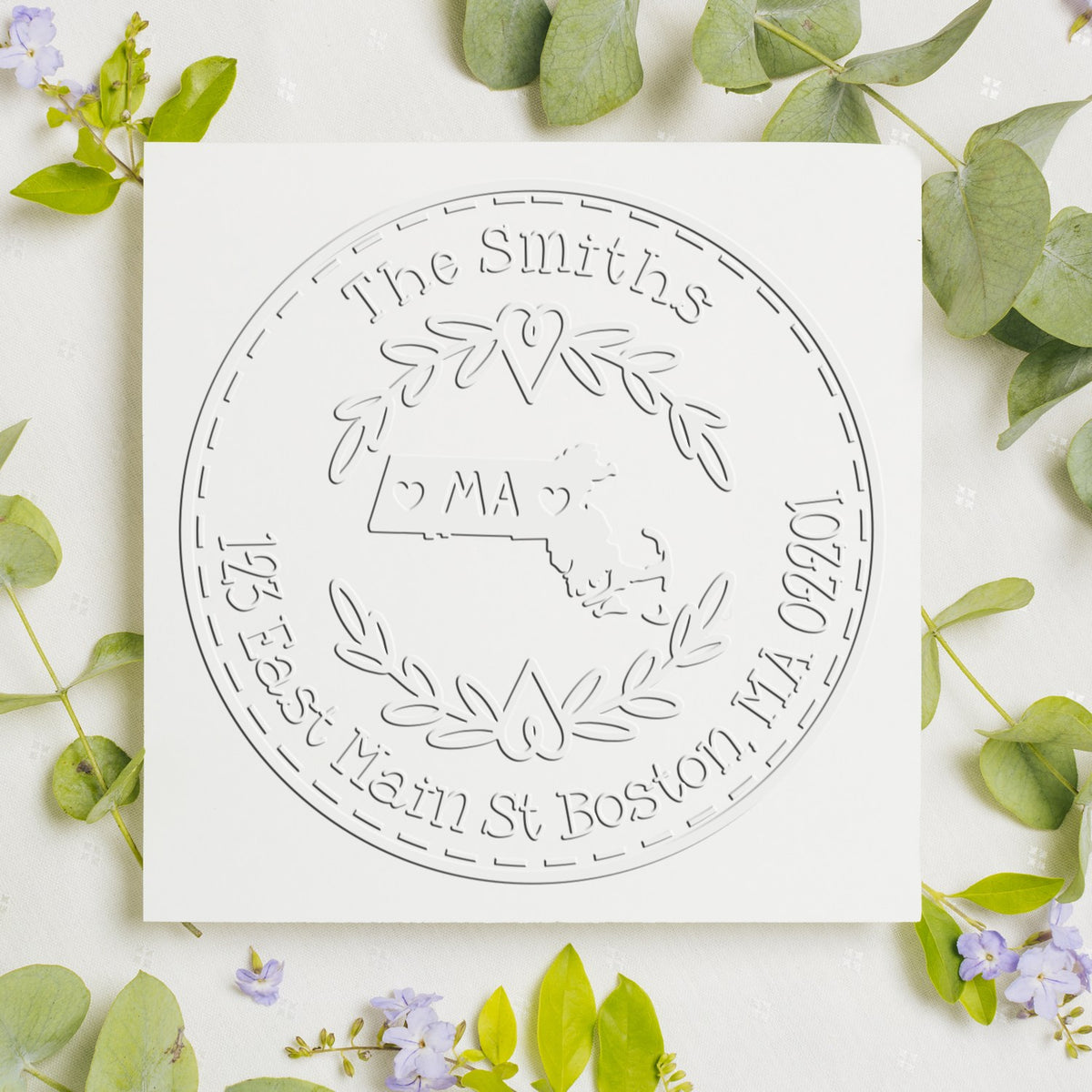 Embossed white custom seal stamp with floral wreath, Massachusetts map, and The Smiths text on a white card, surrounded by green leaves.
