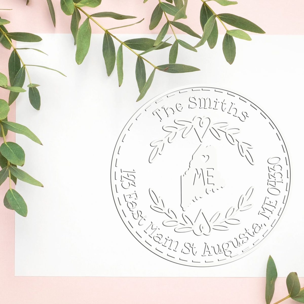 Custom circular stamp seal with engraved family name, silver finish, on white paper with green leaves and pink background.
