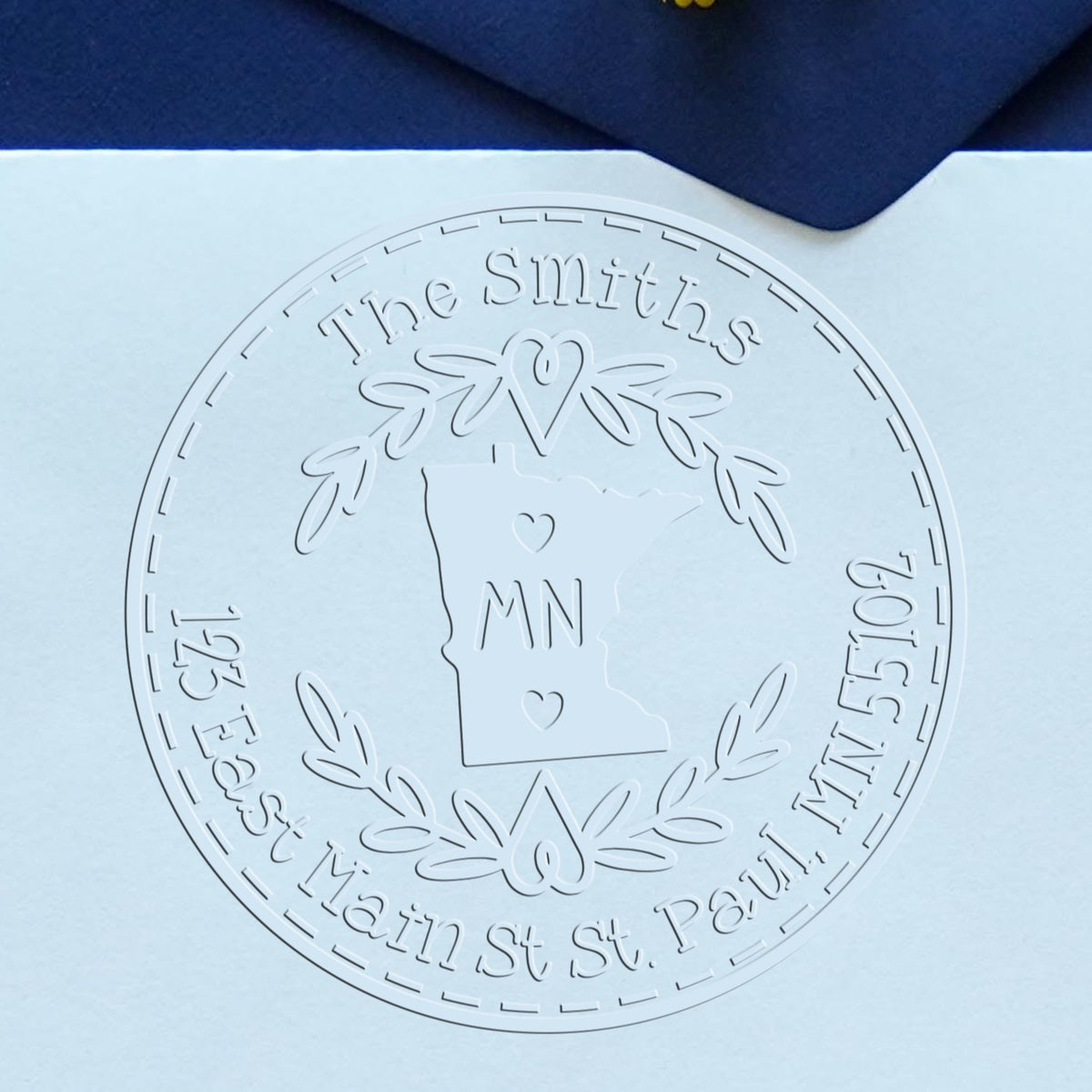 Embossed white custom stamp seal with Minnesota outline and laurel wreath on pale blue paper, framed by dark navy fabric