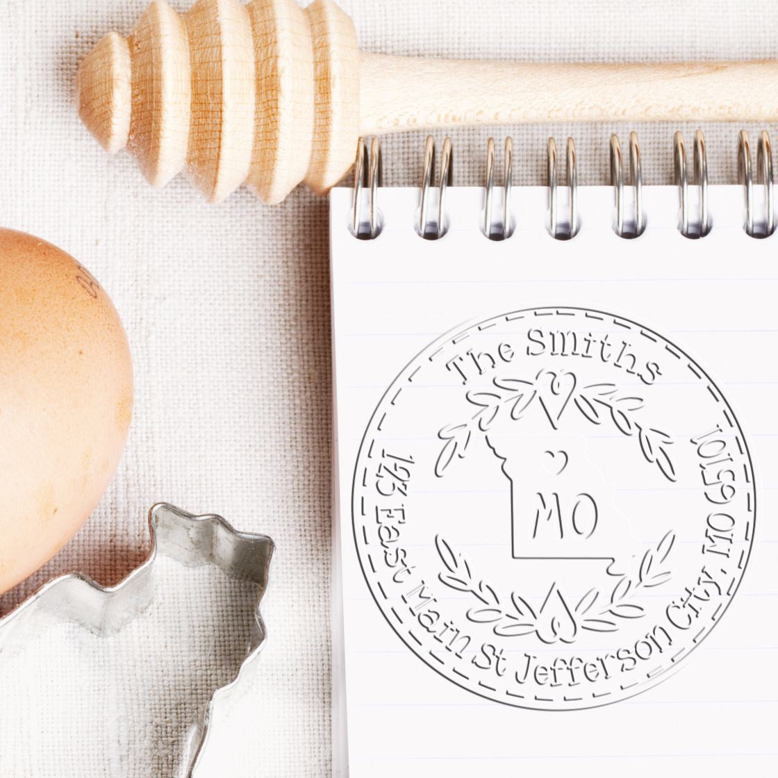 Custom gray Missouri state seal stamp embossed on white spiral notebook, with wooden handle and metal cookie cutter on beige fabric.