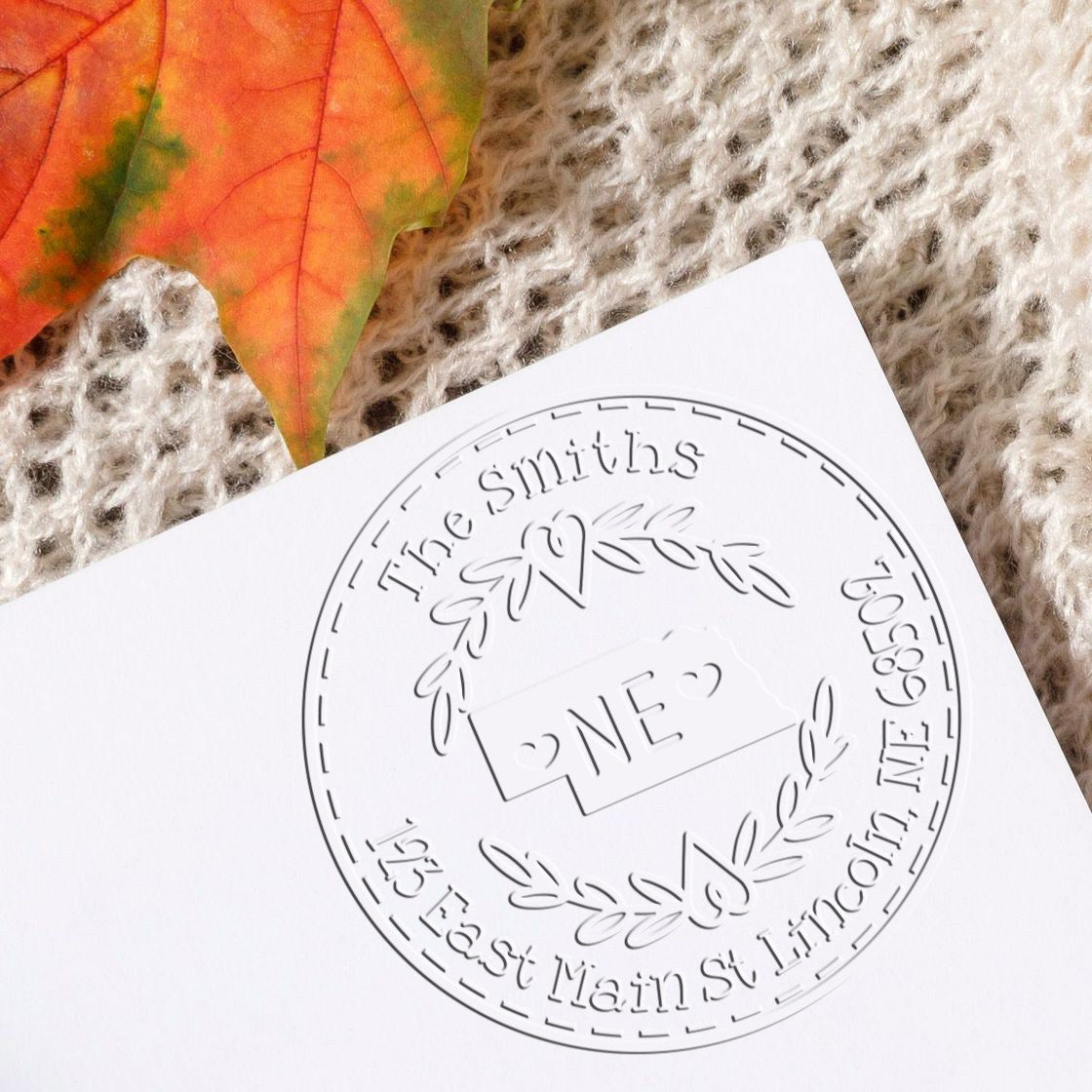 Embossed white custom stamp seal with laurel wreath and The Smiths on white paper, beside an orange autumn leaf.