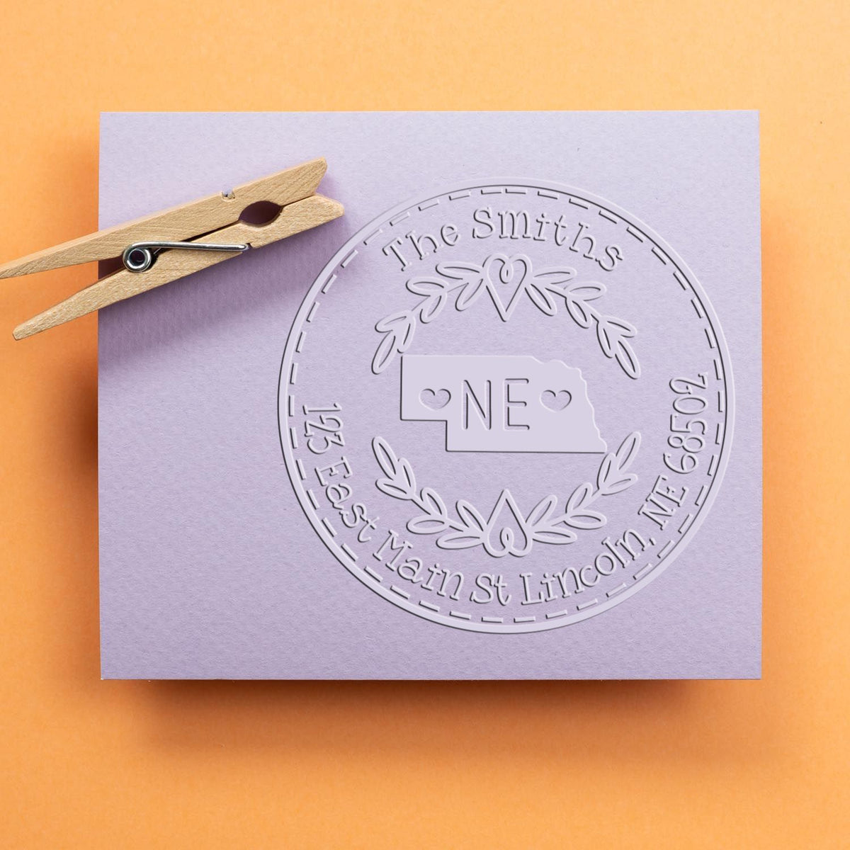 Wooden clothespin beside lilac paper with embossed circular stamp seal for The Smiths and Nebraska outline on orange background.