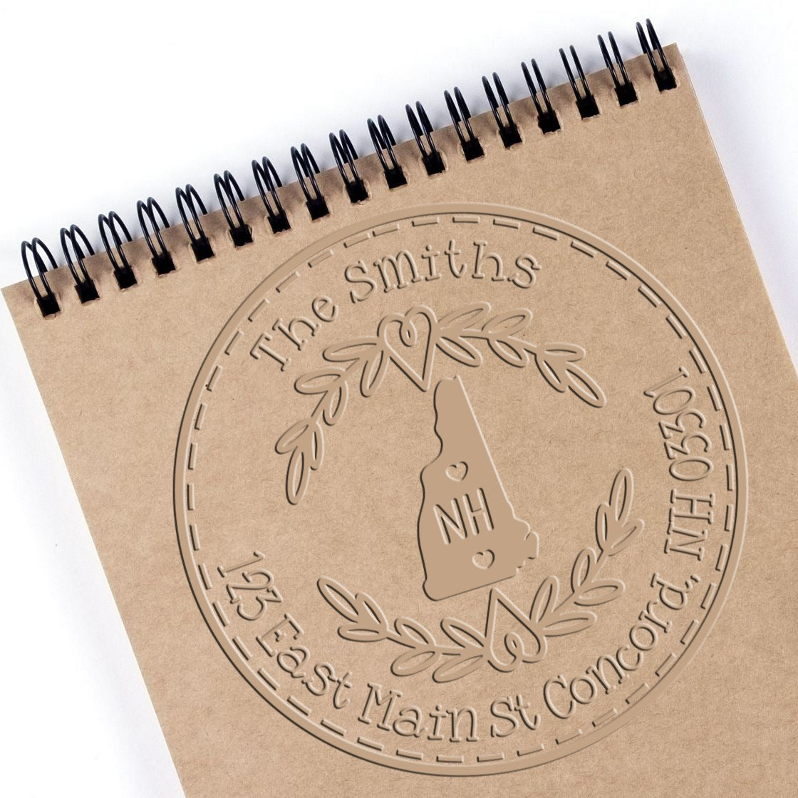Embossed tan custom stamp seal on a spiral notebook, with New Hampshire outline, laurel wreath, and black coil binding.