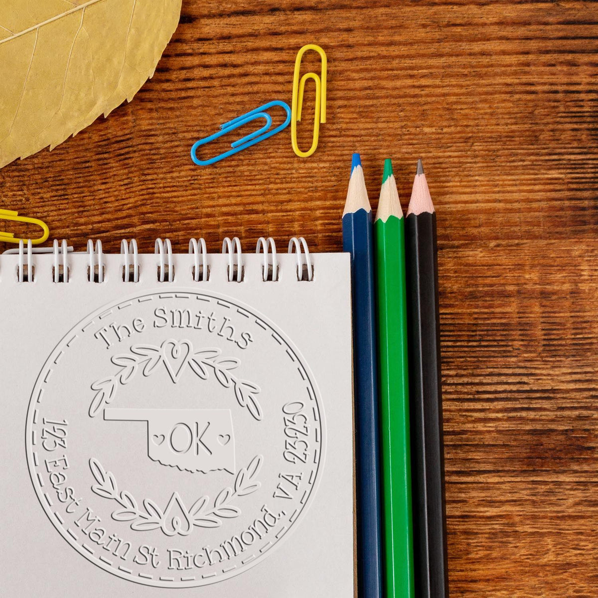 Embossed custom stamp and seal on spiral notepad beside blue, green and black pencils, paper clips, and wood desk.