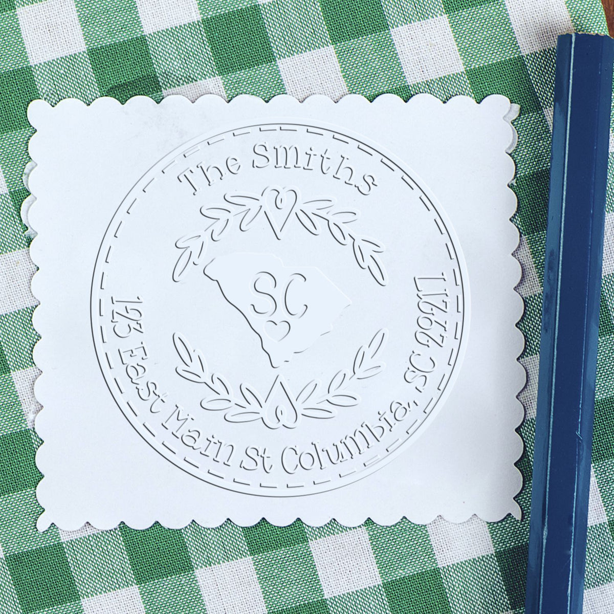 White scalloped stamp and seal sample with embossed circular South Carolina crest on green gingham, beside a blue pencil.