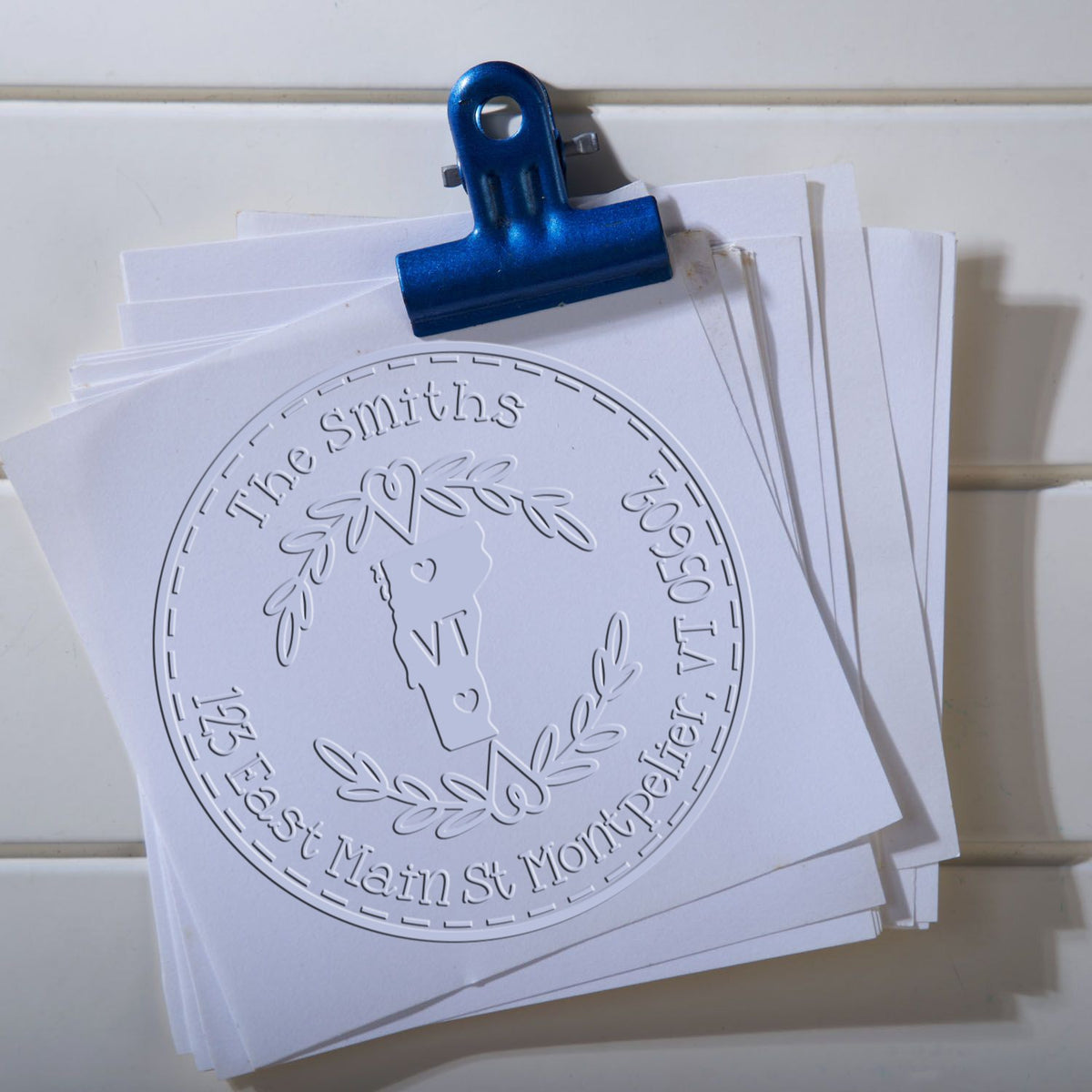 Blue binder clip holding white papers with embossed custom seal stamp reading The Smiths, 195 East Main St Montpelier VT.