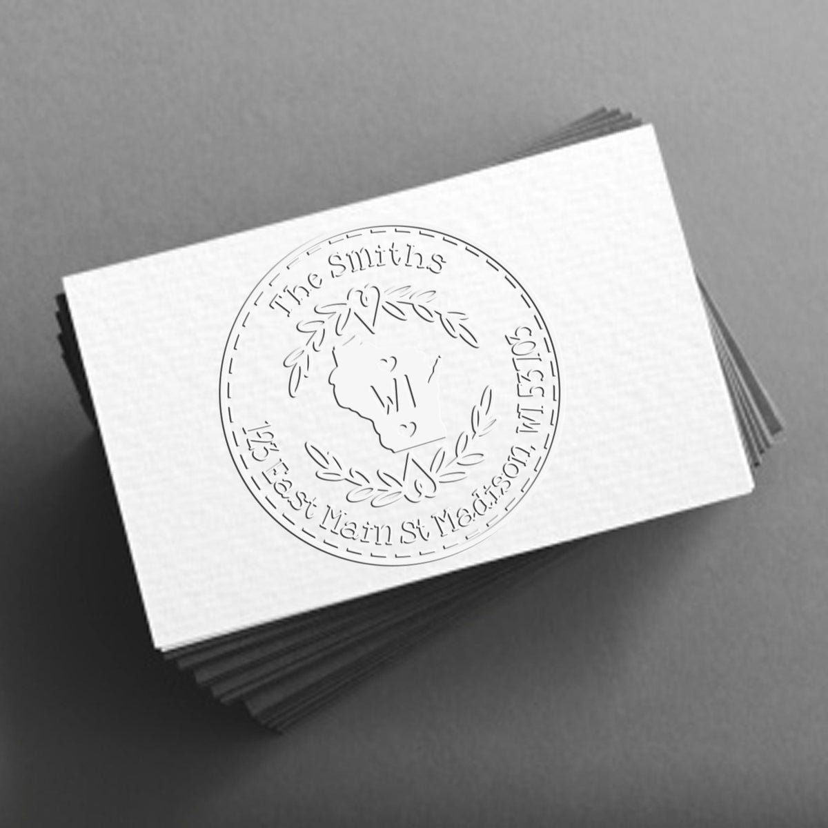 White embossed custom stamp and seal on stacked cards with a circular laurel logo, set against a gray background.