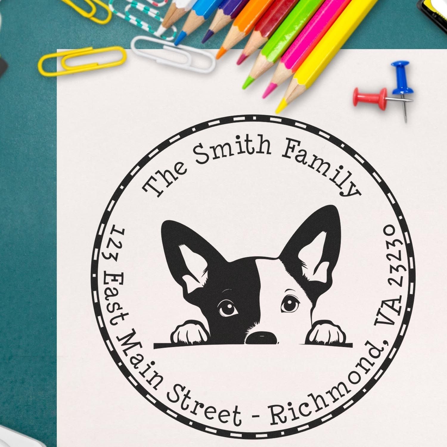 Custom black family stamp seal with dog logo on white paper, surrounded by colorful pencils, paper clips, and push pins on teal.