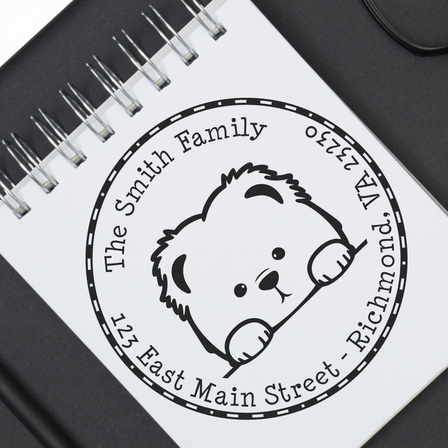 Custom black round stamp seal on a white spiral notebook, featuring a teddy bear and personalized family name and address.