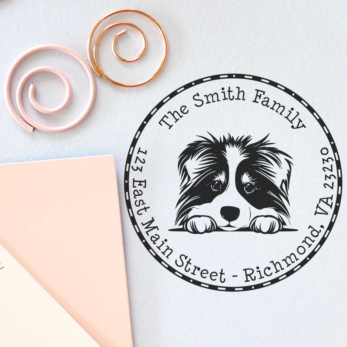 Custom black family address stamp and seal with raccoon design, beside pink and gold spiral embossing tools on white paper
