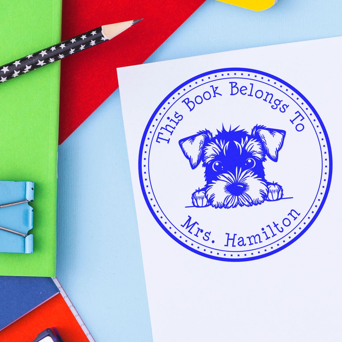 Blue bookplate stamp and seal with dog and This Book Belongs To Mrs. Hamilton, on white paper with colorful notebooks and pencil.