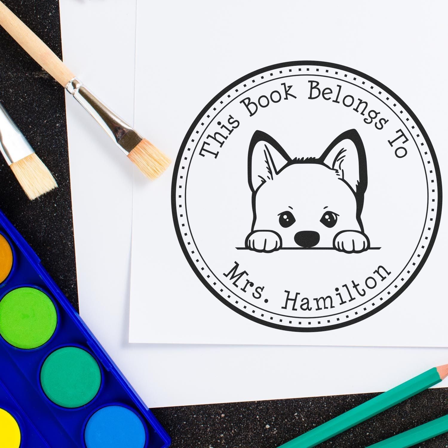 Custom black-and-white round book stamp seal with dog peeking over line, surrounded by paintbrushes and colorful watercolor set.