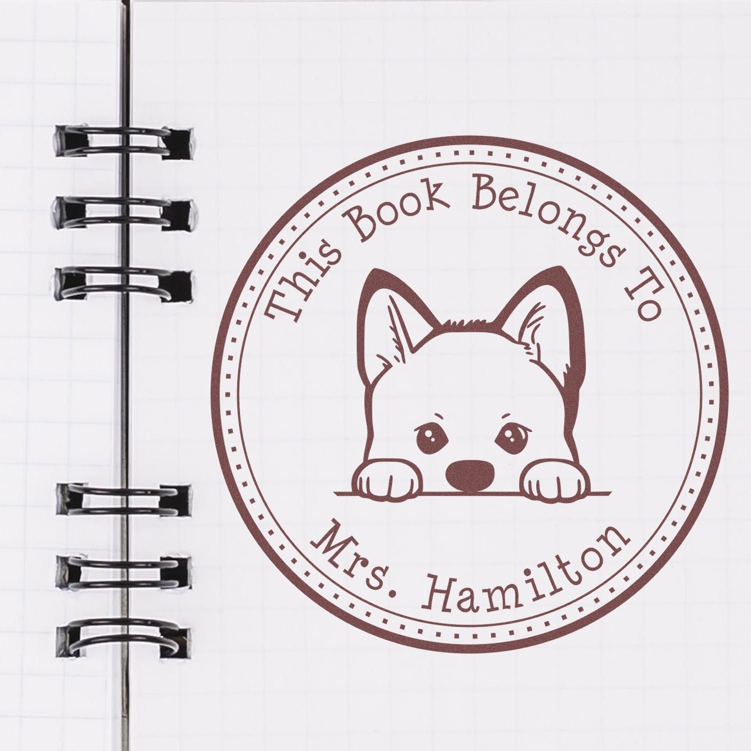 Brown custom book stamp seal with puppy icon and This Book Belongs To Mrs. Hamilton on a notebook page.