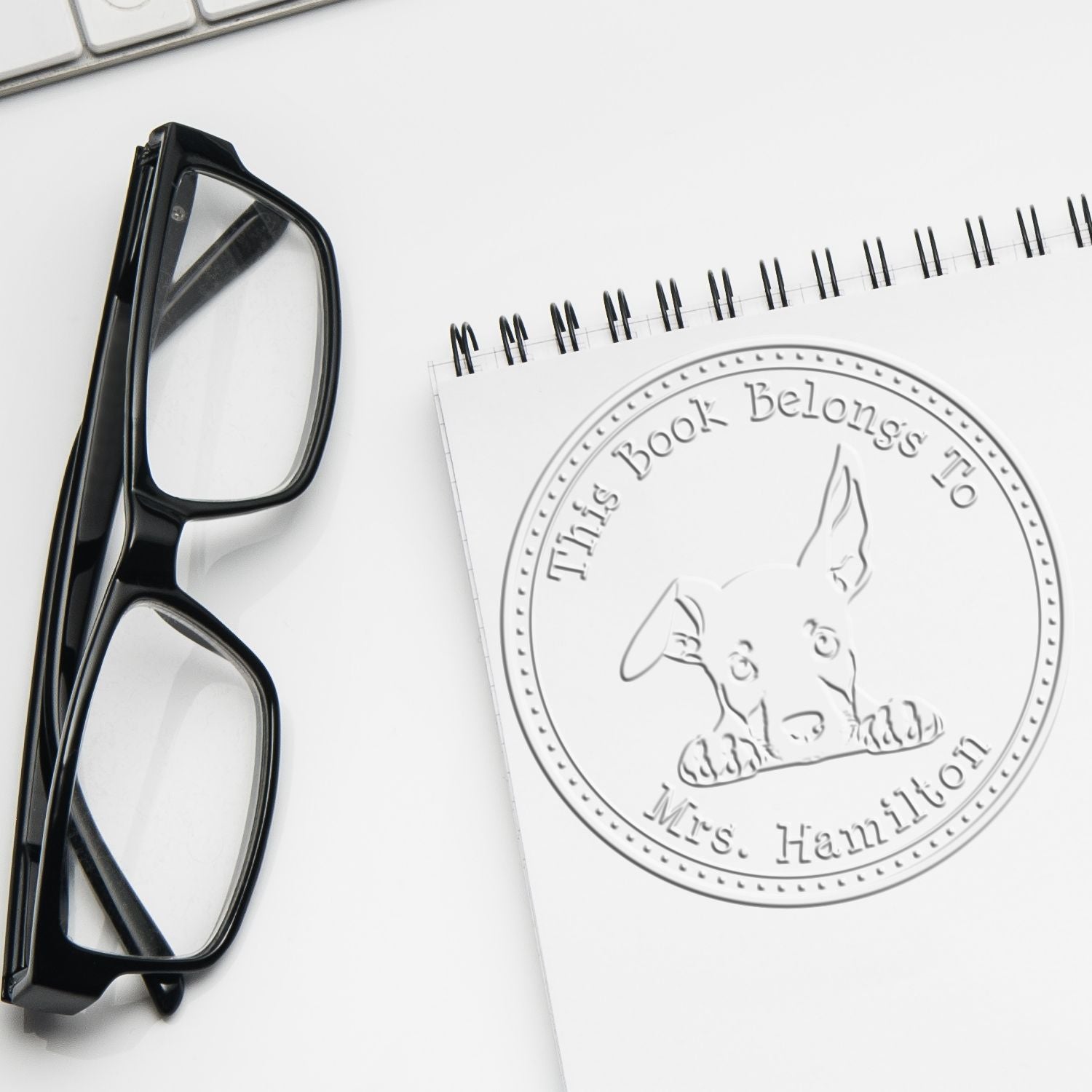 Black glasses beside a white notepad with an embossed custom book stamp seal and dog logo reading This Book Belongs To Mrs. Hamilton
