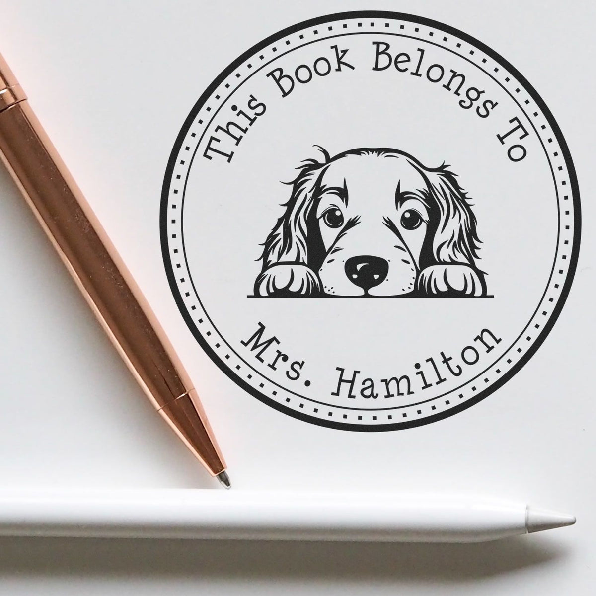 Black custom book seal stamp with dog illustration and Mrs. Hamilton text on white paper, beside a rose-gold pen and white stylus.