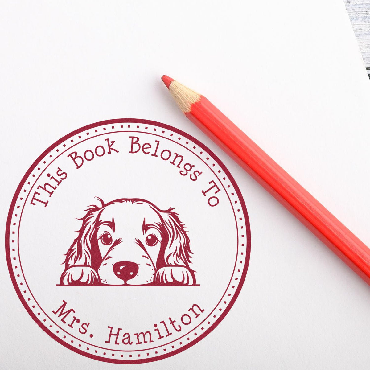 Red custom book ownership stamp and seal with dog illustration, This Book Belongs to Mrs. Hamilton, beside a red pencil.