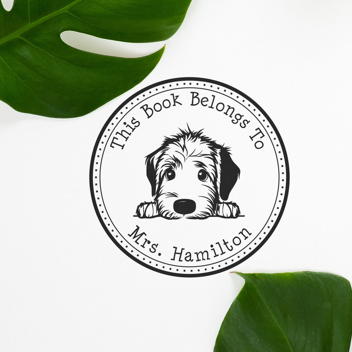 Circular black custom book stamp seal with dog graphic and This Book Belongs To Mrs. Hamilton on white, framed by green leaves.
