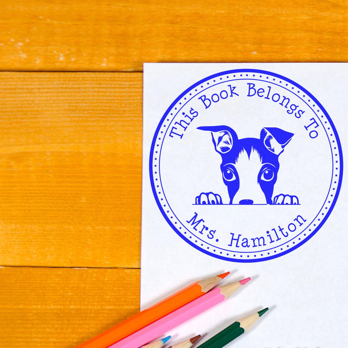 Blue custom book ownership stamp seal on white paper with dog mascot, on orange wood tabletop, with colored pencils.