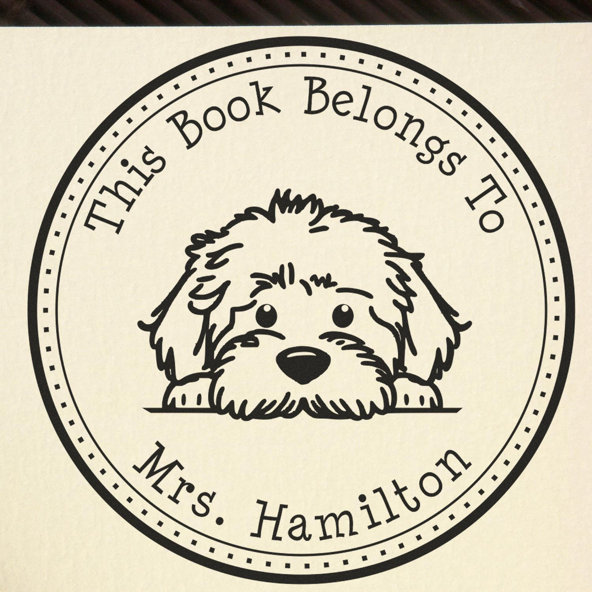 Black custom book stamp seal with fluffy dog illustration and text This Book Belongs To Mrs. Hamilton on cream background.