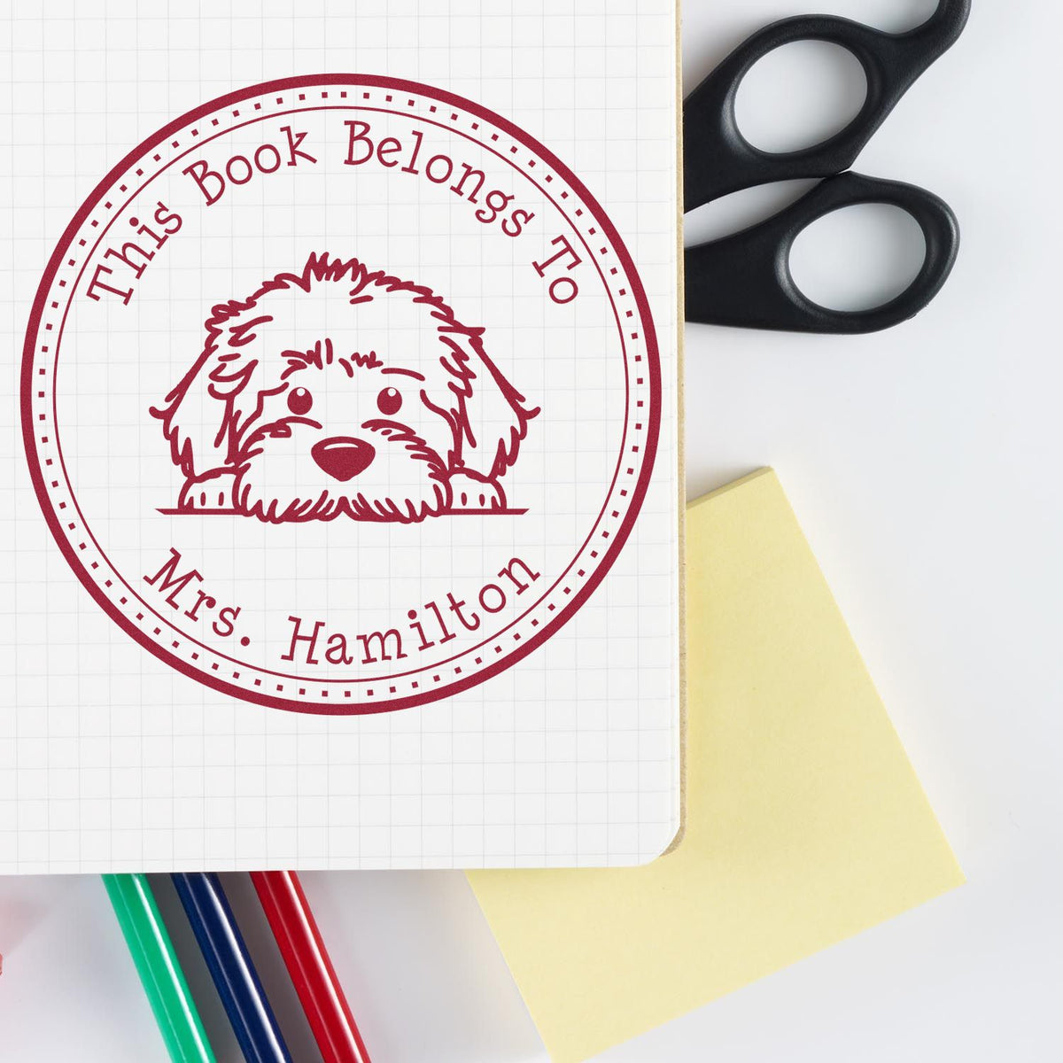 Maroon This Book Belongs to Mrs. Hamilton dog stamp seal on white grid paper, with scissors, sticky note, and pens.