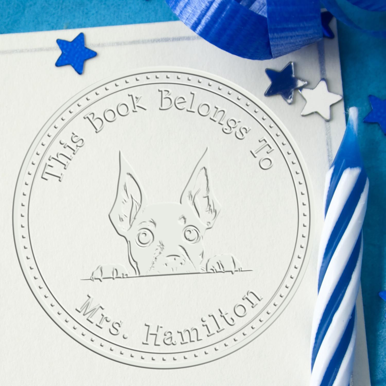 Embossed book seal with peeking dog, blue stars, and ribbon; circular stamp reads This Book Belongs To Mrs. Hamilton.