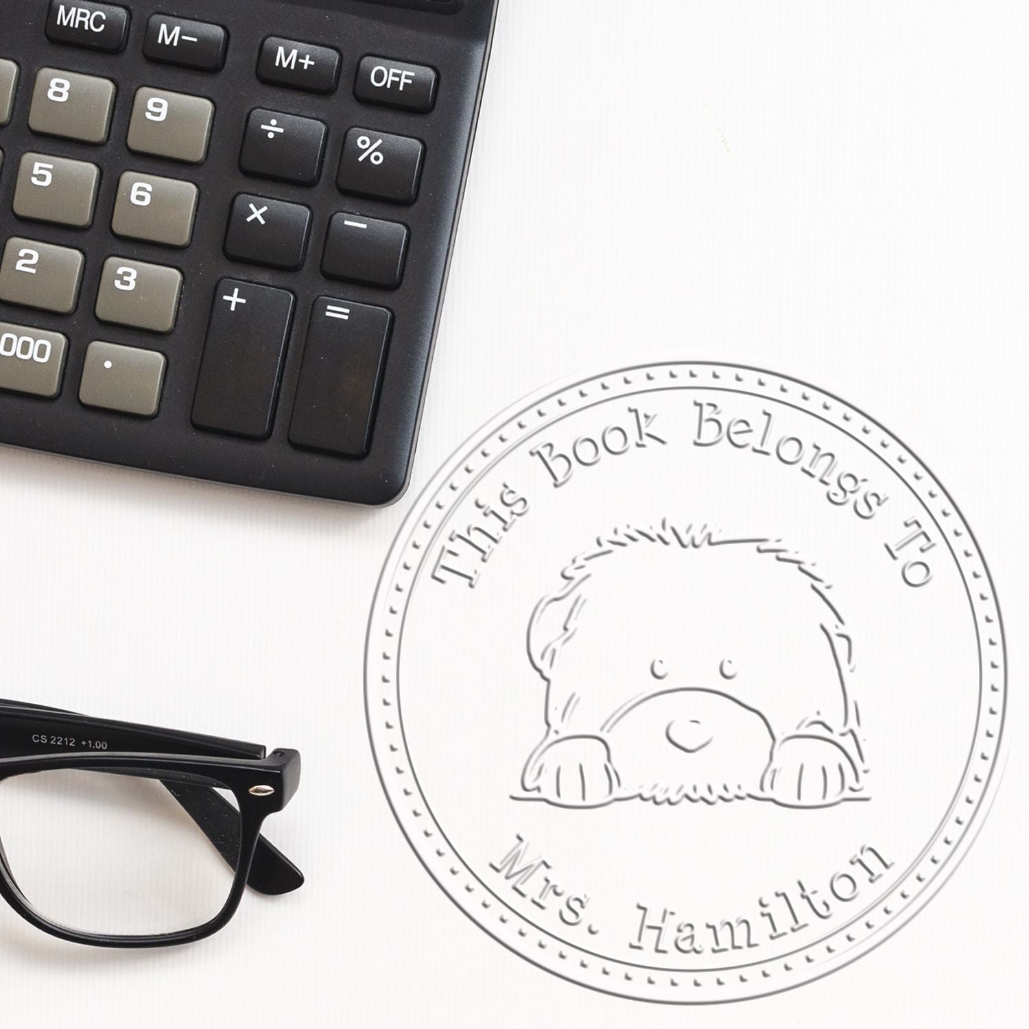 Round custom book stamp seal with dog design and This Book Belongs to Mrs. Hamilton on white, beside black calculator and glasses.