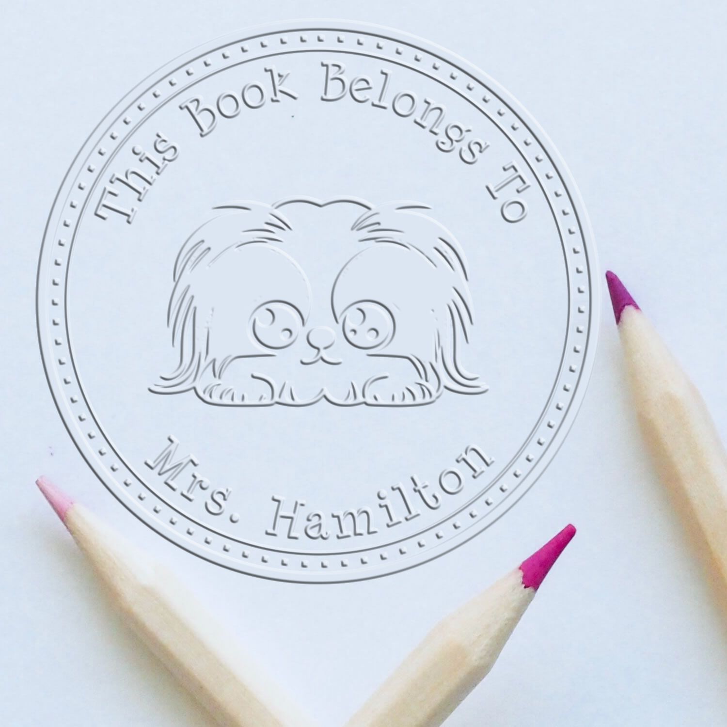 White embossed circular book stamp and seal with dog icon and This Book Belongs To Mrs. Hamilton text on pale paper.