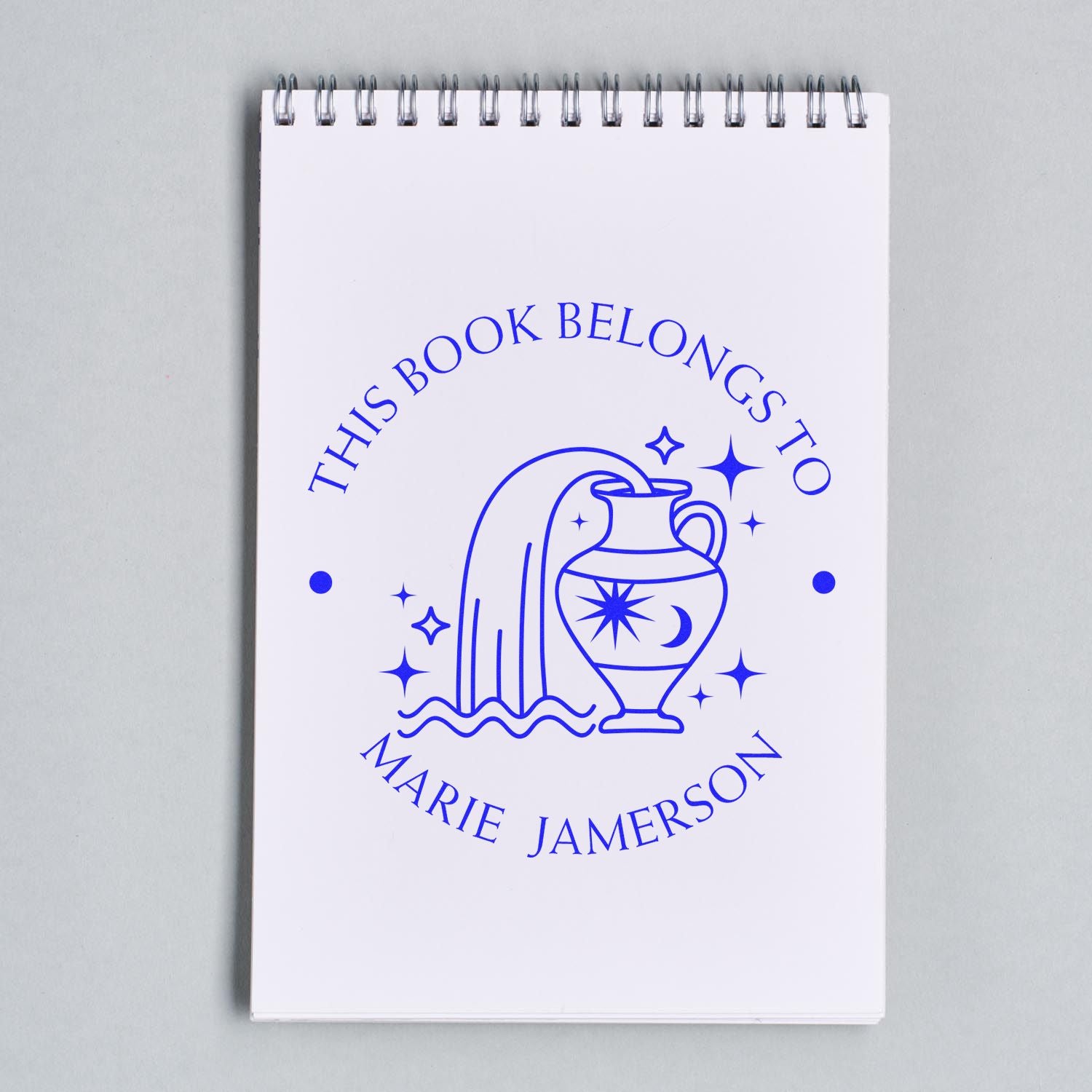 Blue ex libris stamp seal on a white spiral notebook, with vase, stars, and text This Book Belongs to Marie Jamerson.