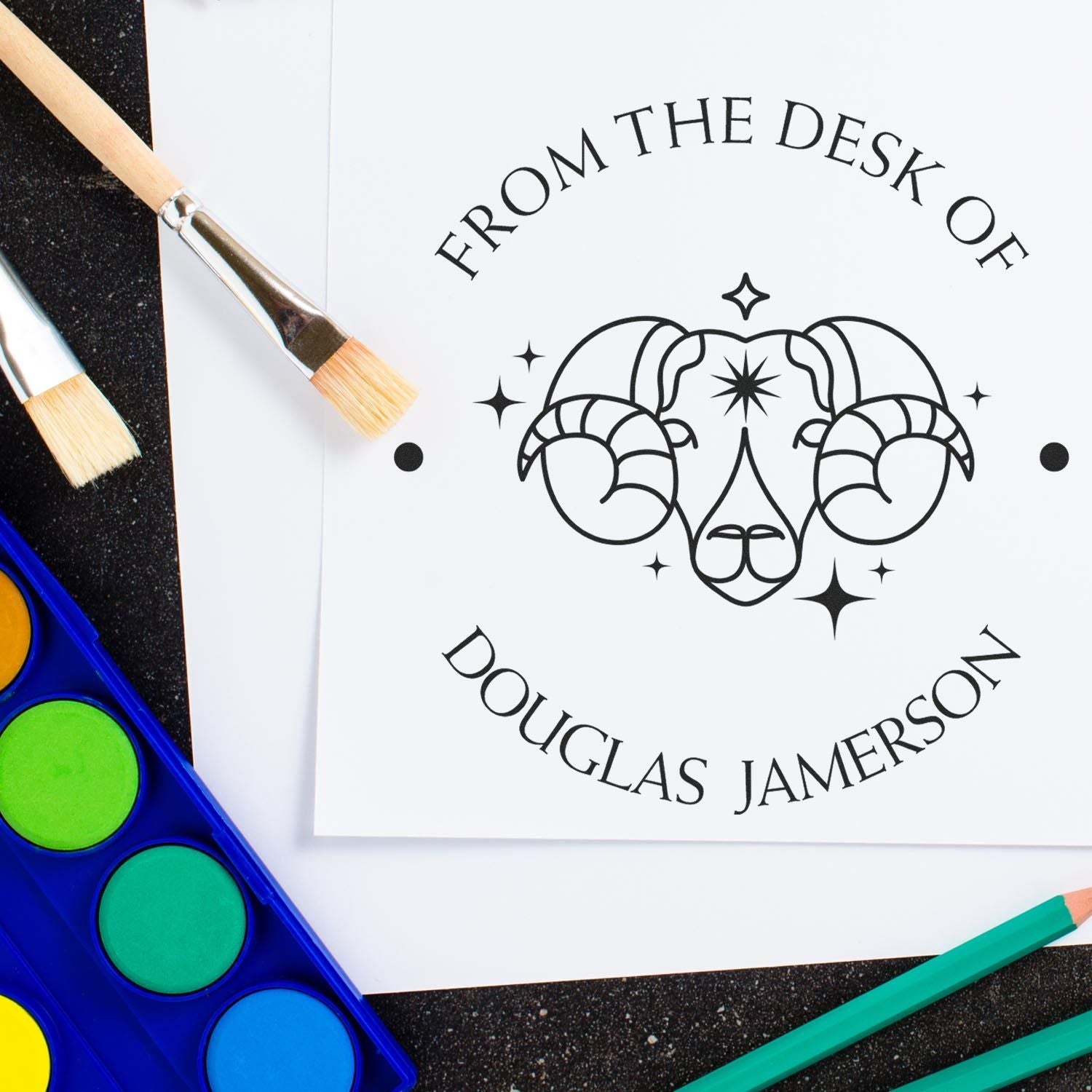 Custom stamp and seal products with black ram logo on white paper, plus paint brushes, watercolor palette, and teal pencils.