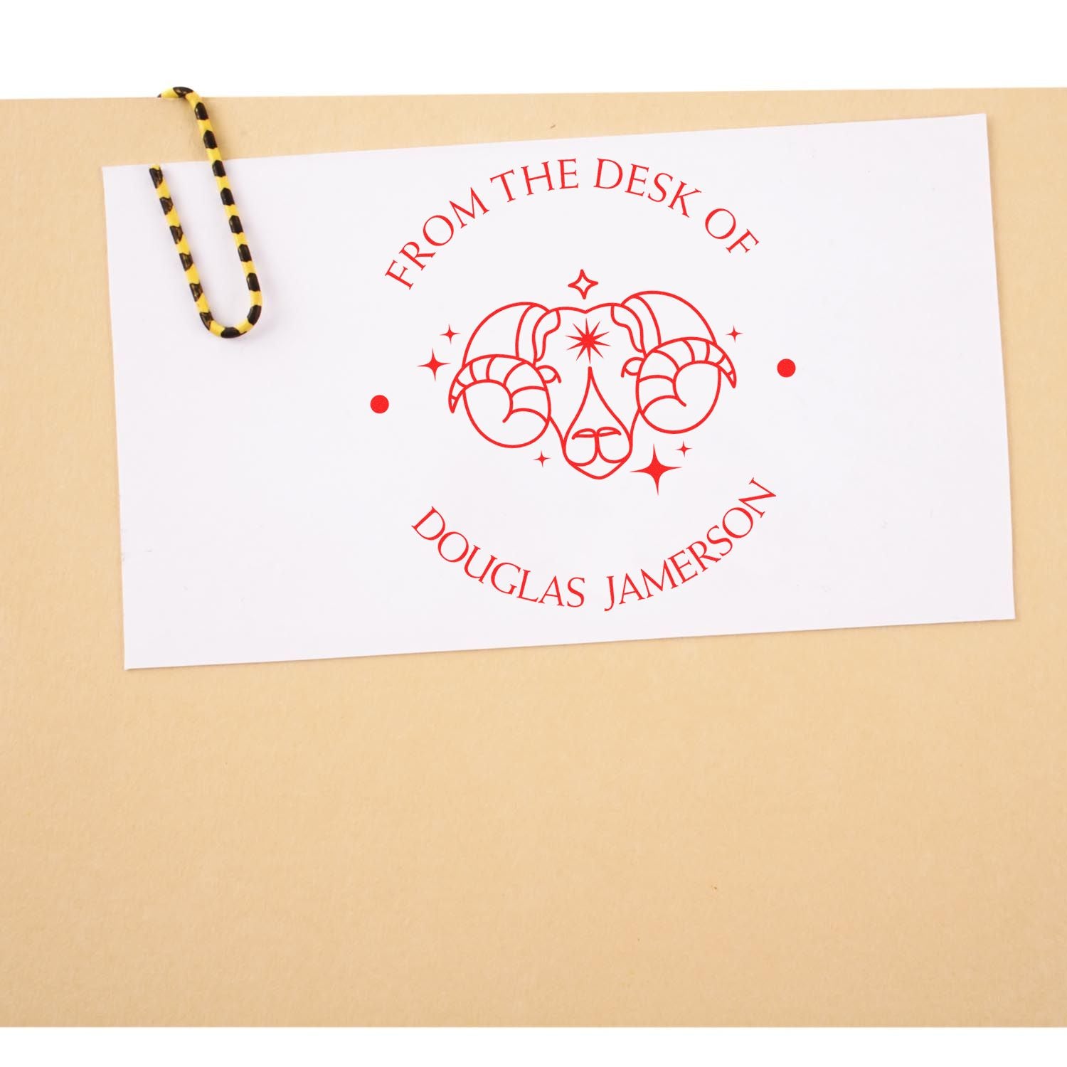 Red custom desk stamp and seal with ram logo on white card, clipped by black-and-yellow binder clip against tan background.