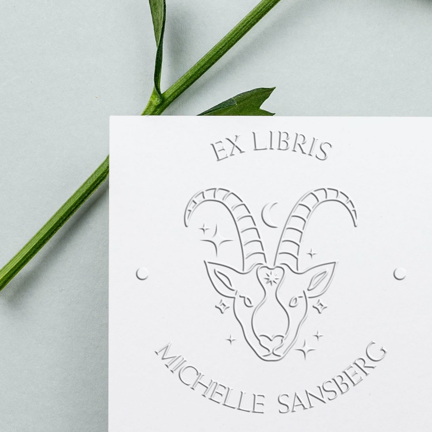 White embossed ex libris stamp seal with ram emblem and Michelle Sansberg text on pale background, crossed by a green stem.