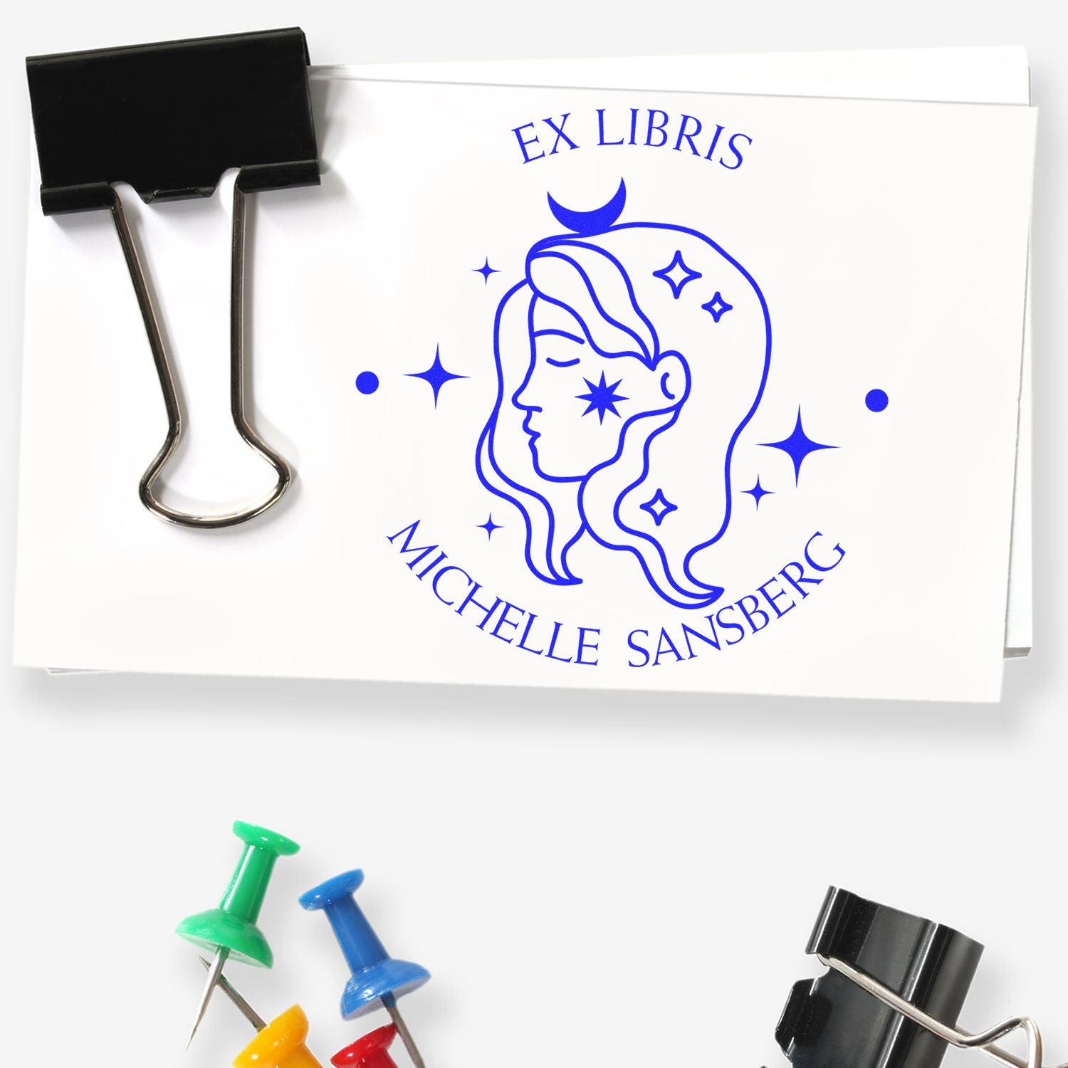 Blue custom ex libris stamp and seal on white paper, with Michelle Sansberg profile, black binder clip and colorful push pins.