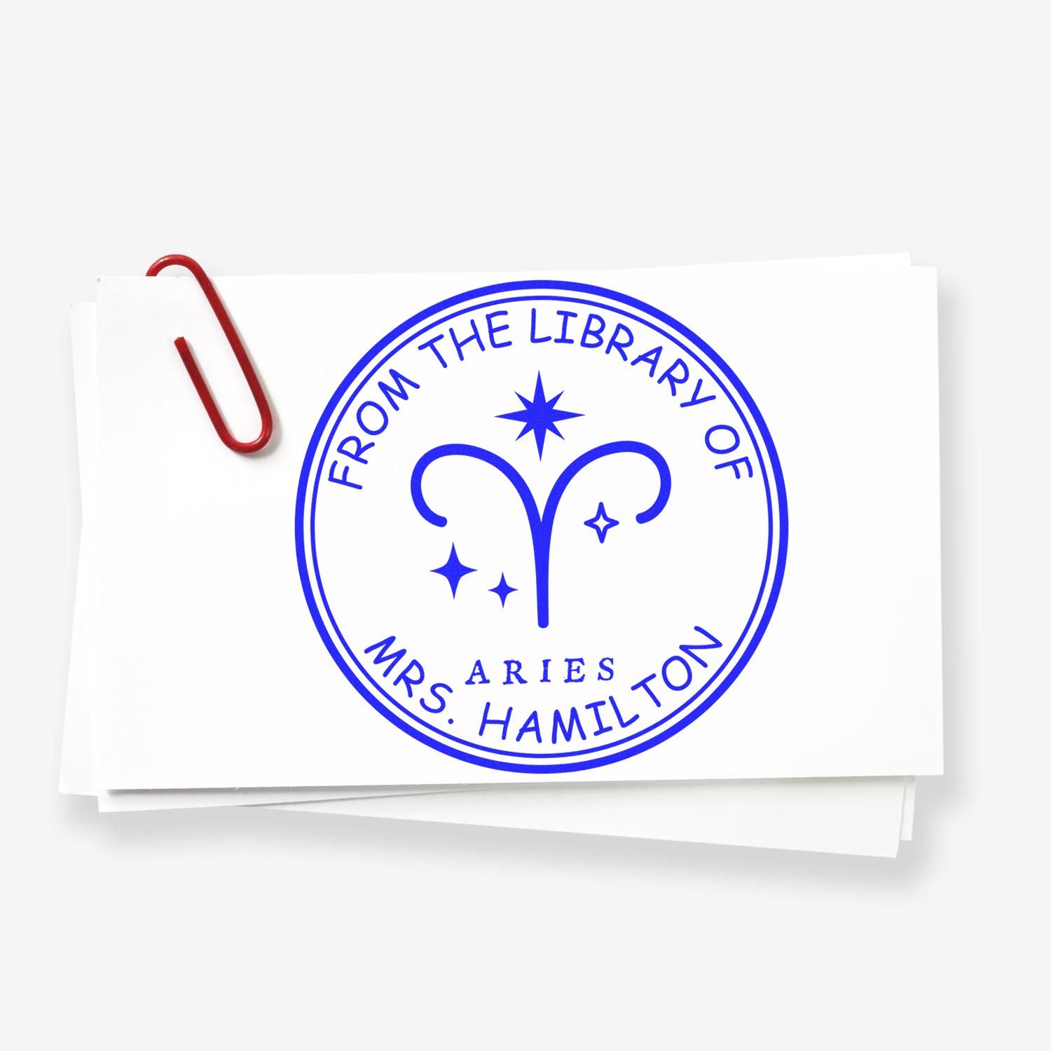 Blue custom library stamp seal with Aries symbol and From the Library of Mrs. Hamilton on white paper, red paperclip.