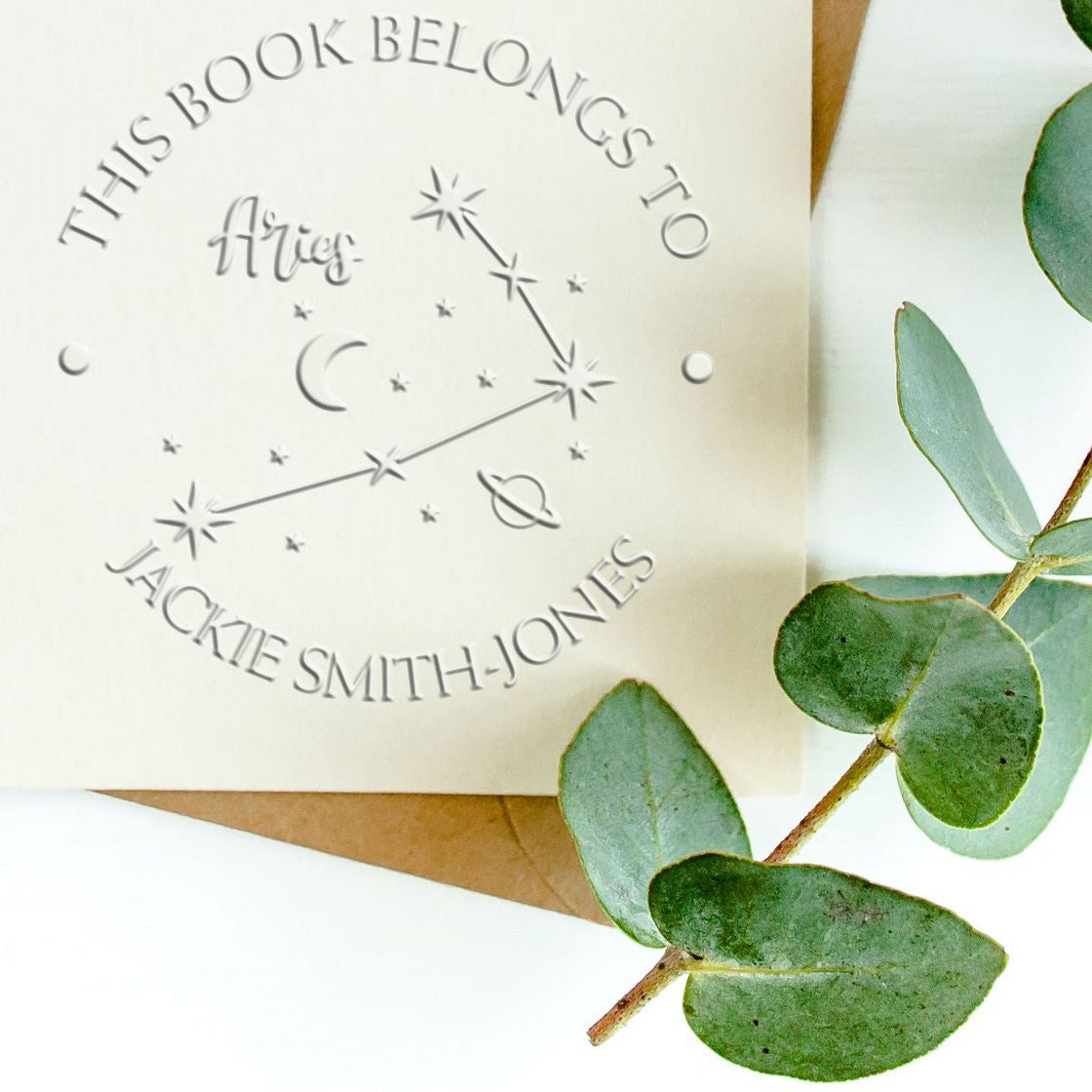 Custom embossed stamp and seal for a bookplate, featuring Aries constellation, silver-gray text, and eucalyptus leaves.