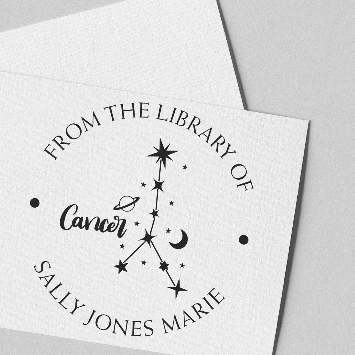 Black custom library stamp seal on white textured paper, featuring Cancer zodiac art, stars, moon, and arched name text.