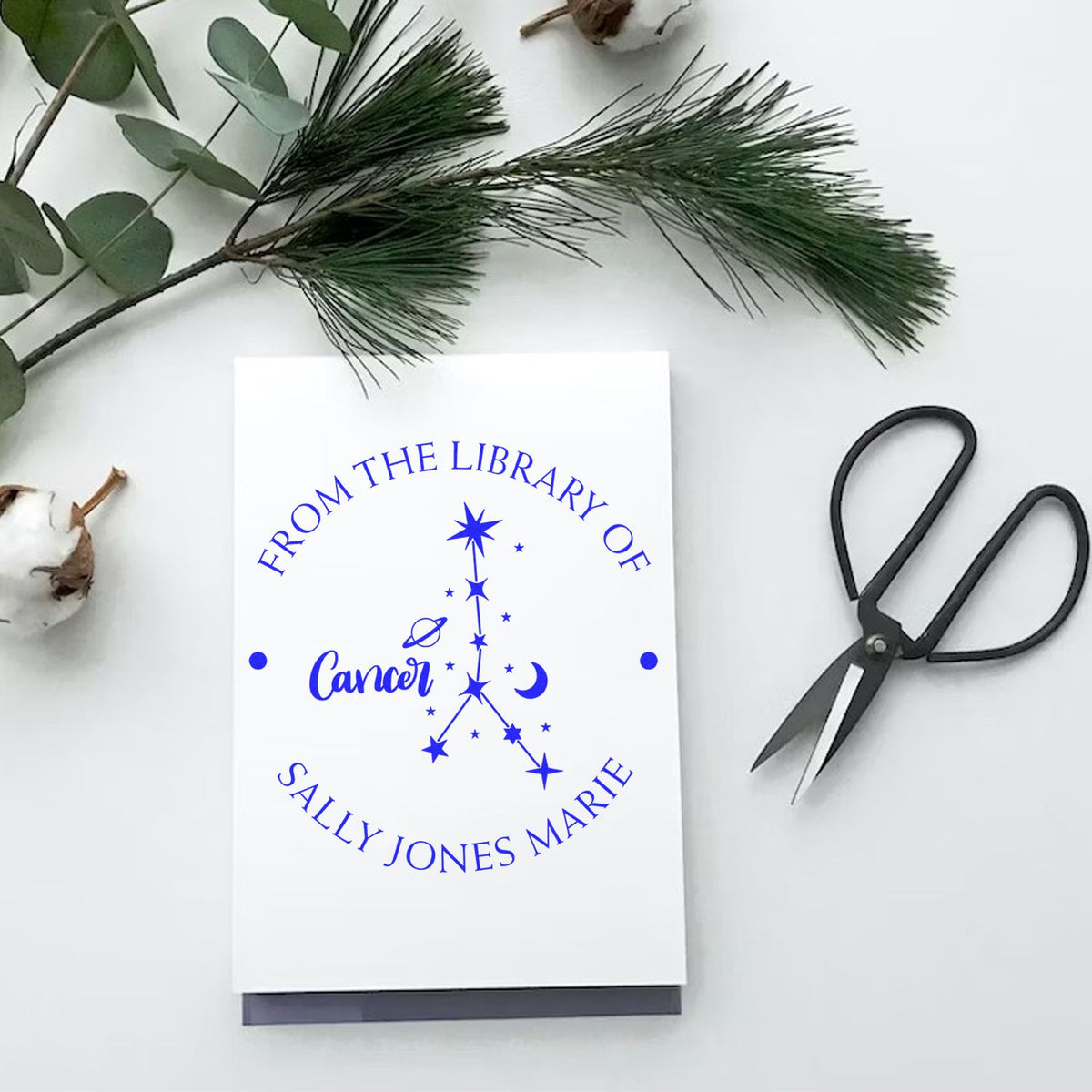 White custom library stamp seal with blue constellation design and name, beside black scissors and greenery.