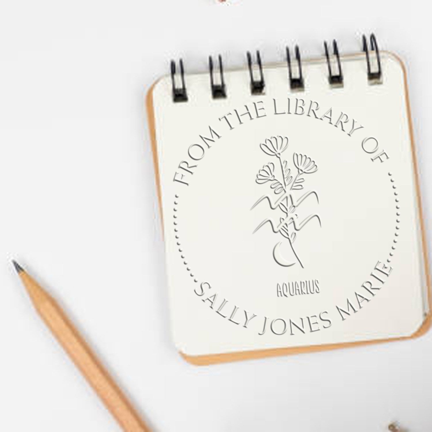 Cream spiral notebook with embossed Aquarius library stamp seal, floral motif, tan edges, and black binding on a white background.