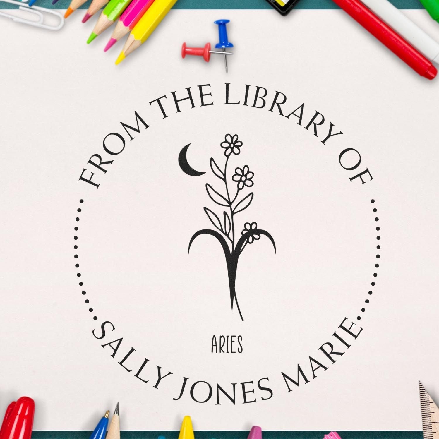 Black personalized library stamp seal with Aries floral emblem on white, framed by colorful pencils and push pins.