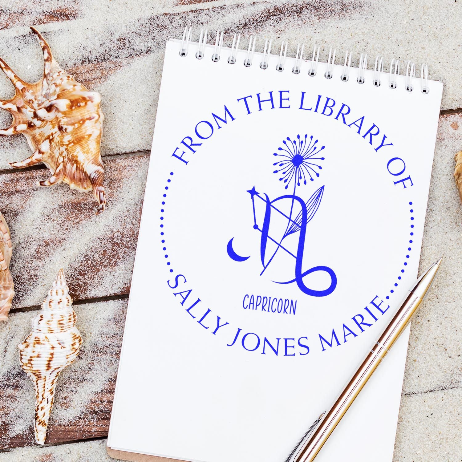 Blue Capricorn custom book stamp and seal on white spiral notebook, with seashells and gold pen on sandy background.
