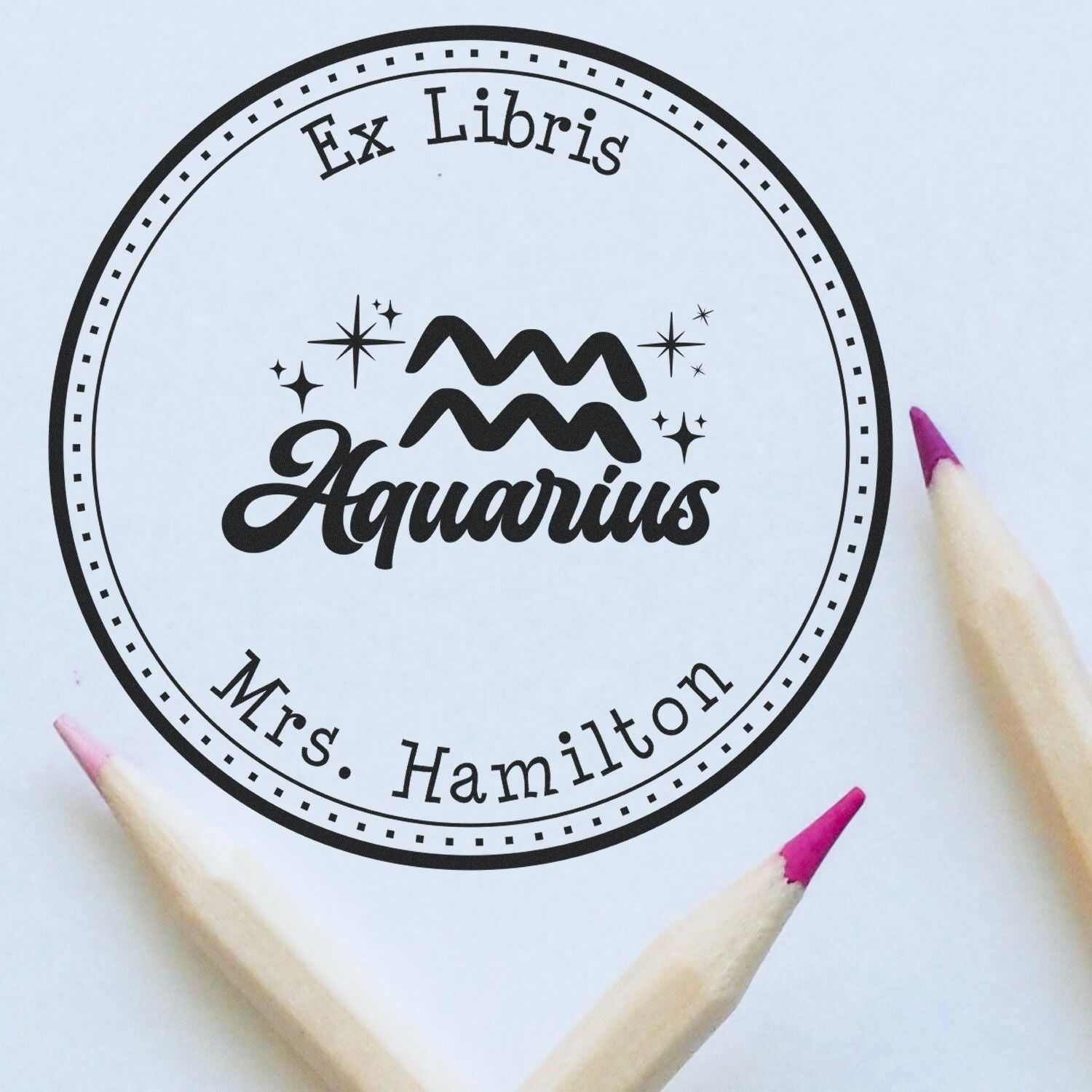 Black circular ex libris Aquarius stamp seal with Mrs. Hamilton text and stars, shown with purple pencil tips on a white background.