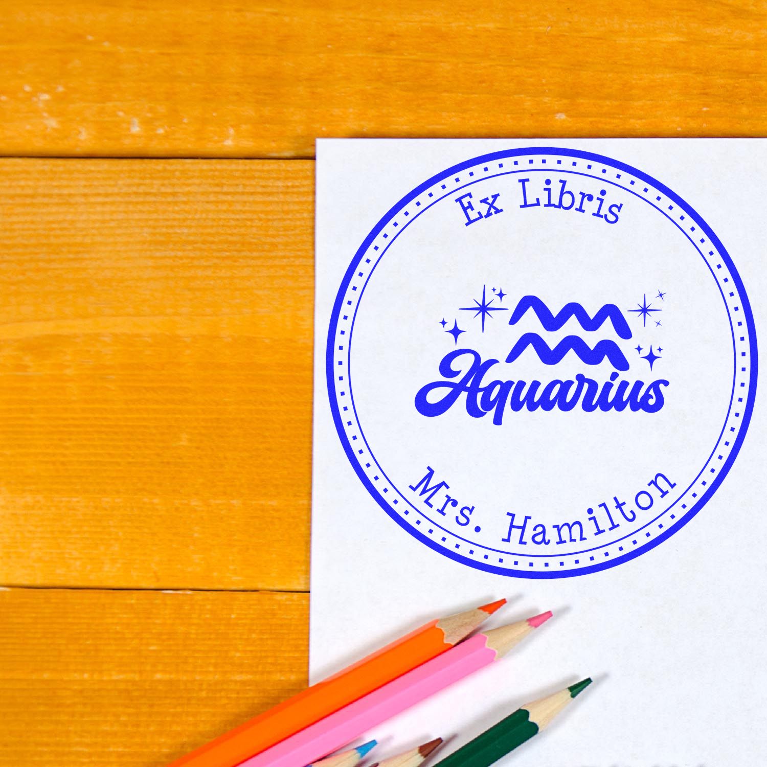 Blue Aquarius ex libris stamp seal for Mrs. Hamilton on white paper, with colored pencils on a warm wood table.