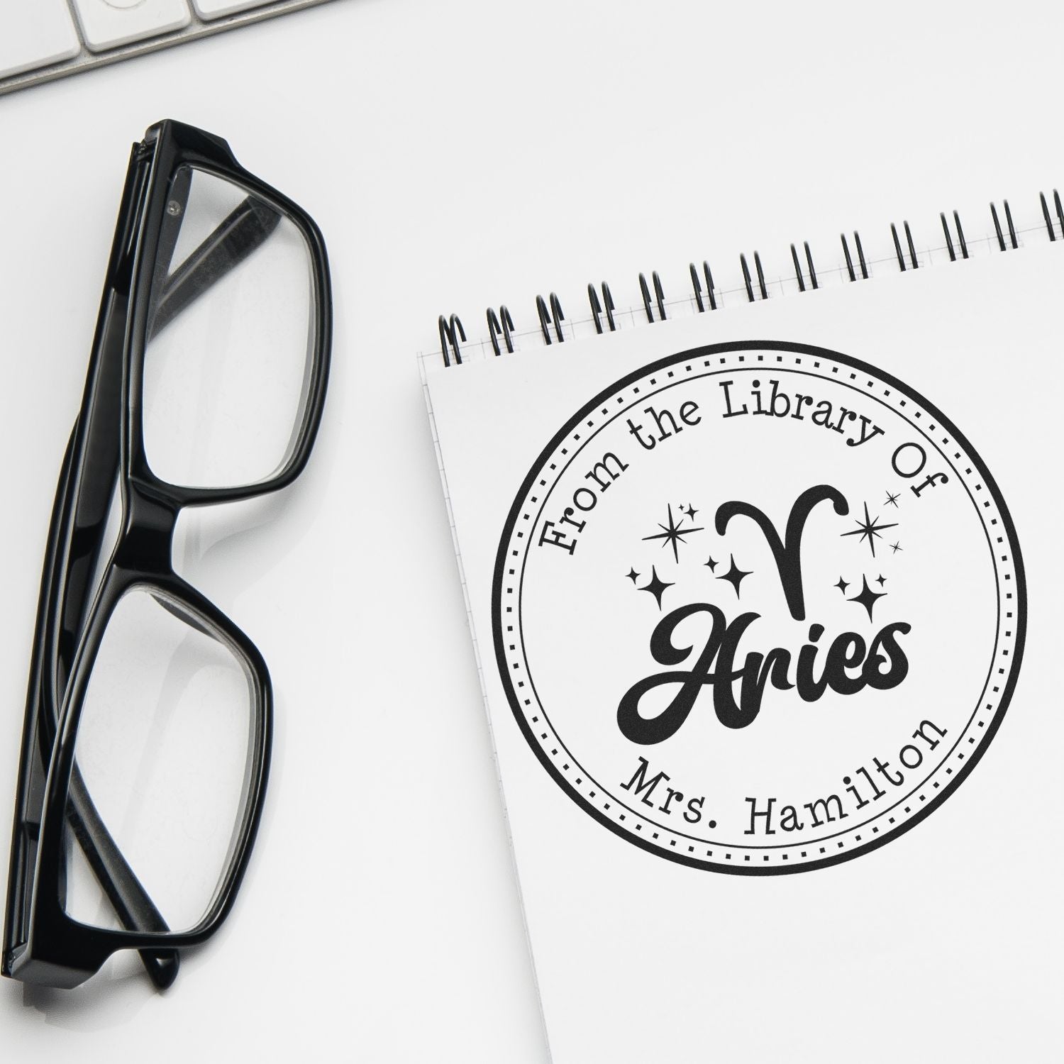 Black custom library stamp and seal beside black eyeglasses on a white notebook with Aries "Mrs. Hamilton" design.