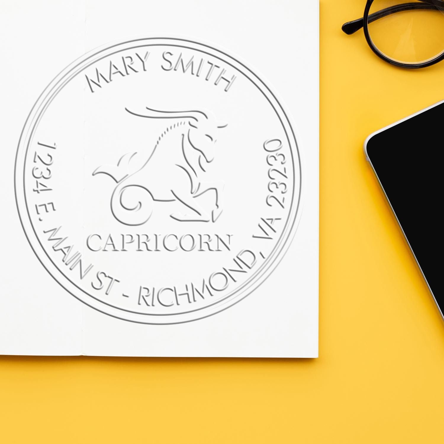 Custom silver Capricorn stamp seal for Mary Smith on white paper, with yellow background, glasses and smartphone edge.