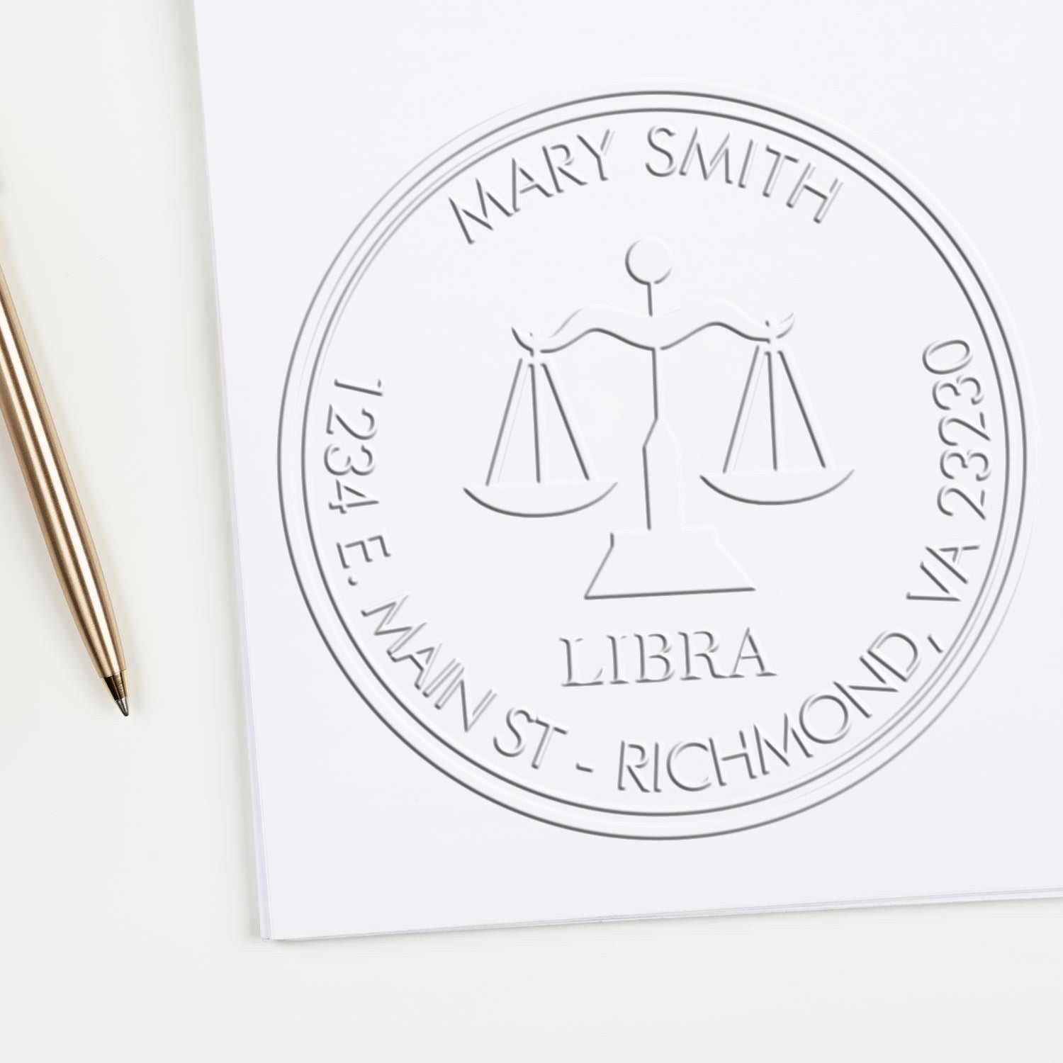 Embossed silver legal seal stamp with scales of justice, Mary Smith, Libra, and address on white paper beside a gold pen.